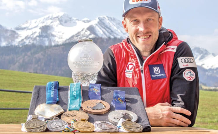 Ein Mann in Sportkleidung sitzt vor einer alpinen Kulisse. Vor ihm liegen mehrere Medaillen und eine große Glastrophäe auf einem Tisch.