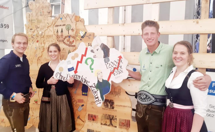 Vier junge Erwachsene in traditioneller bayerischer Kleidung stehen lächelnd vor einer Holzwand mit kreativen Karten- und Grafiken-Elementen.