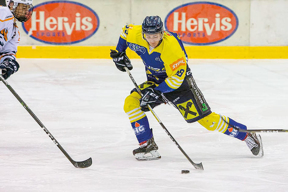 Ein Eishockeyspieler im gelben Trikot bewegt den Puck auf dem Eis, während ein Gegenspieler versucht, ihn zu blockieren. Im Hintergrund sind Werbetafeln zu sehen.