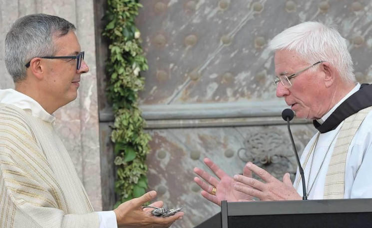 Zwei Geistliche in religiösen Gewändern stehen sich gegenüber. Einer hält eine Schale, während der andere spricht, möglicherweise bei einer Zeremonie.