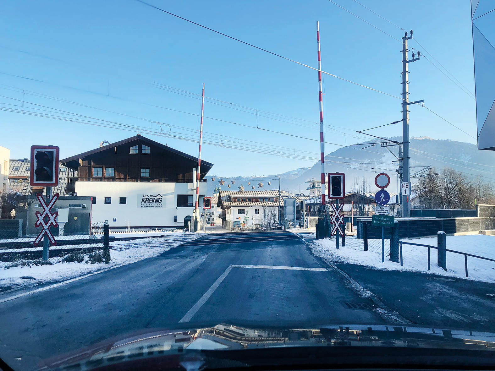 Ein verschneiter Bahnübergang mit Schranken und Signalen, umgeben von Gebäuden im Alpenstil und einem Berg im Hintergrund an einem klaren Tag.