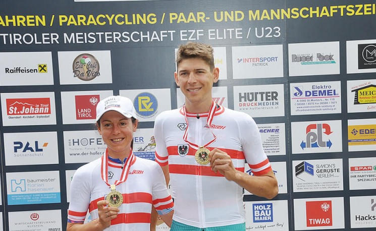 Zwei Radsportler in Rennkleidung stehen vor einer Sponsorentafel und halten Medaillen. Sie lächeln und tragen ein Trikot mit roten Streifen.