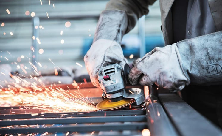 Eine Person schleift Metall mit einem Winkelschleifer, Funken fliegen. Sie trägt schützende Handschuhe und arbeitet in einer Werkstattumgebung.
