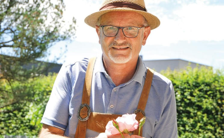 Ein älterer Mann mit Brille und Strohhut lächelt im Freien und hält eine rosa Blume. Er trägt ein blaues Hemd mit braunen Hosenträgern.