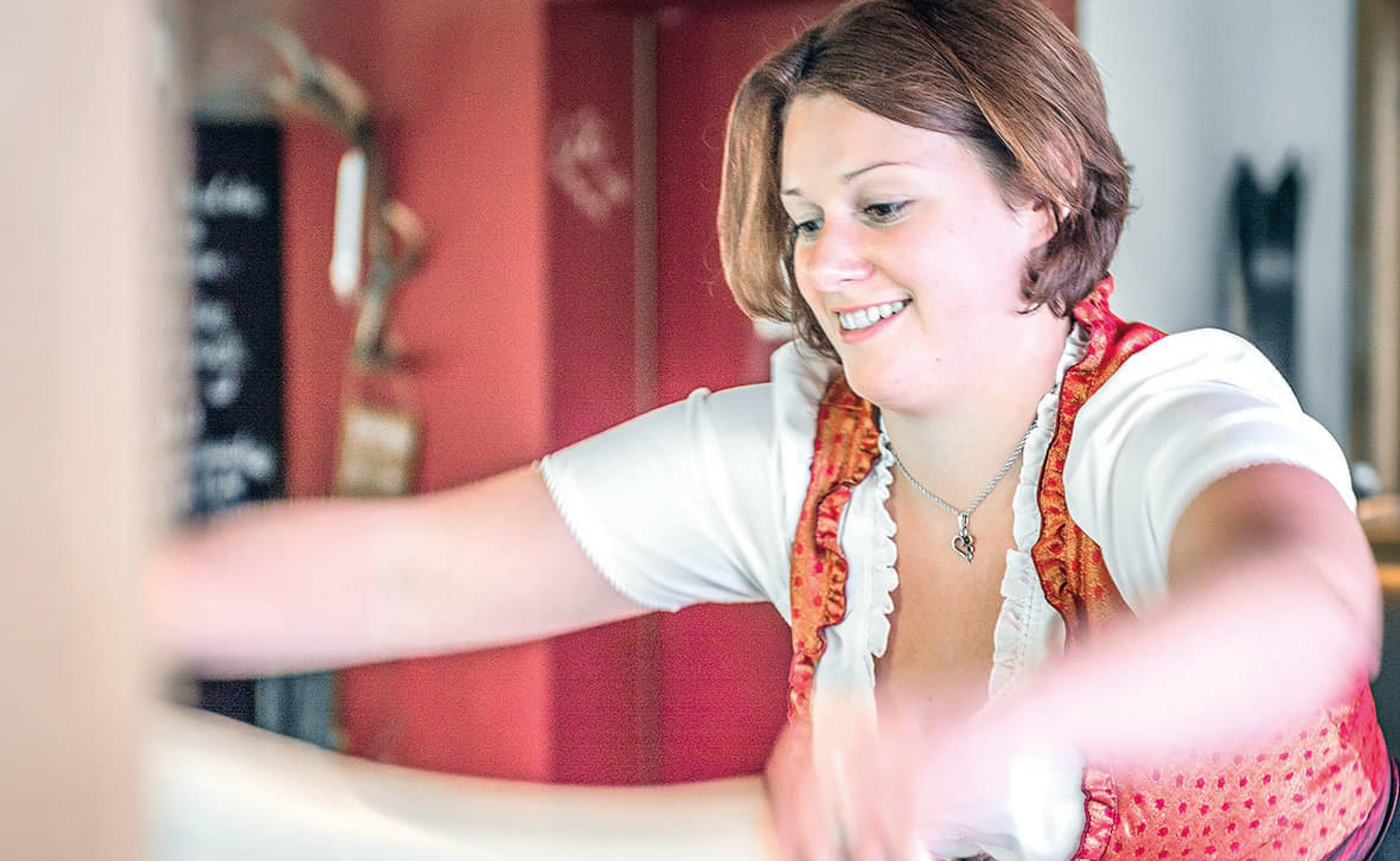 Frau in traditioneller bayerischer Kleidung arbeitet lächelnd in einem hellen Raum mit roten Akzenten.