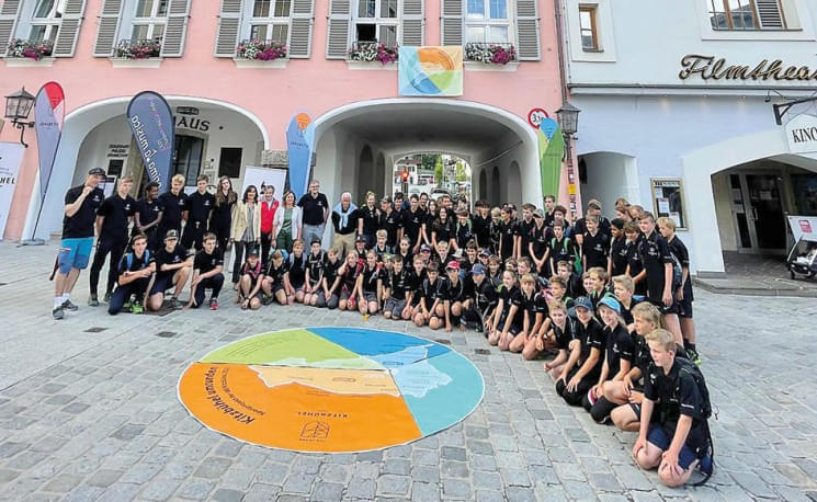 Gruppe junger Menschen in schwarzen T-Shirts posiert in einer Fußgängerzone vor einem bemalten Kreismuster auf dem Pflaster und einem historischen Gebäude.