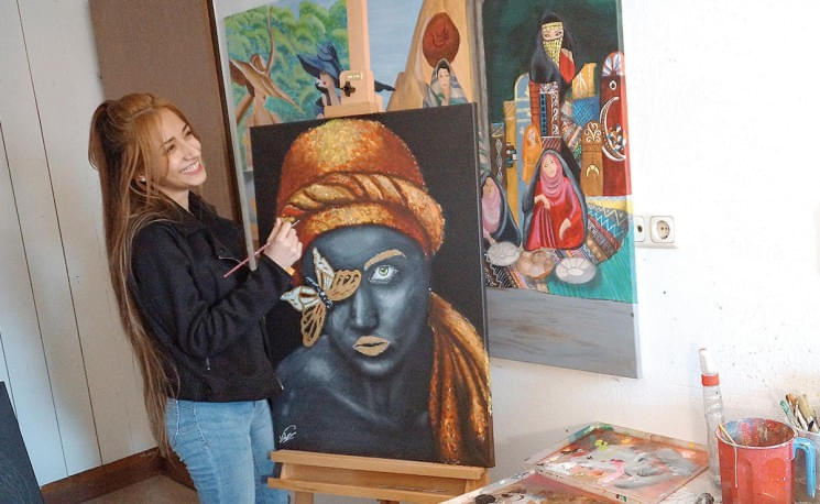 Eine Frau steht lächelnd neben einer Staffelei mit einem Gemälde einer Person mit goldener Kopfbedeckung. Im Hintergrund hängen weitere bunte Kunstwerke.