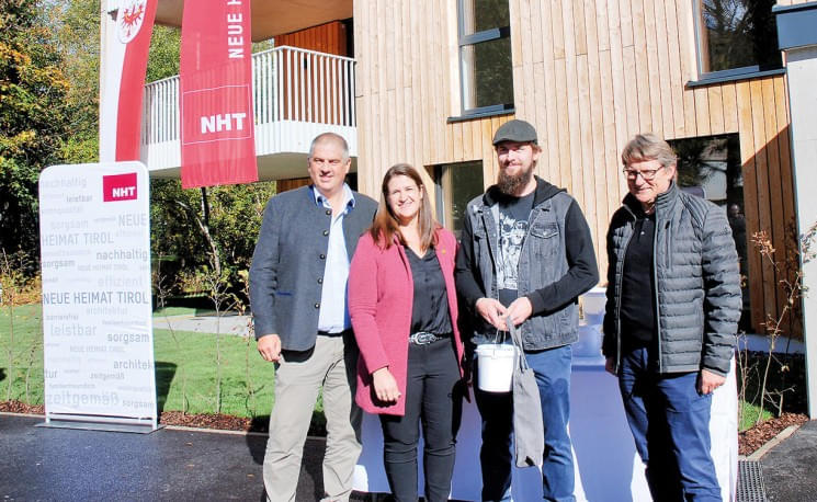 Vier Personen stehen vor einem modernen Holzgebäude mit einem Aufsteller und Banner. Sie lächeln in die Kamera und das Wetter scheint sonnig zu sein.