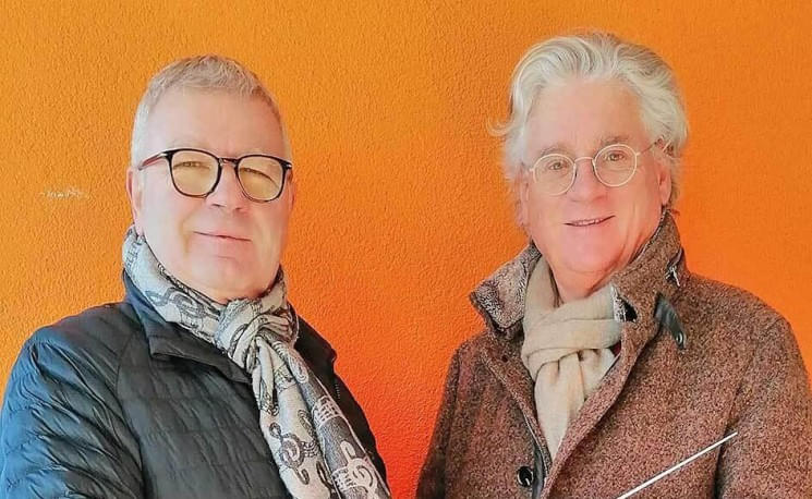 Zwei ältere Männer mit Brillen und Schals stehen vor einer orangen Wand und lächeln in die Kamera.