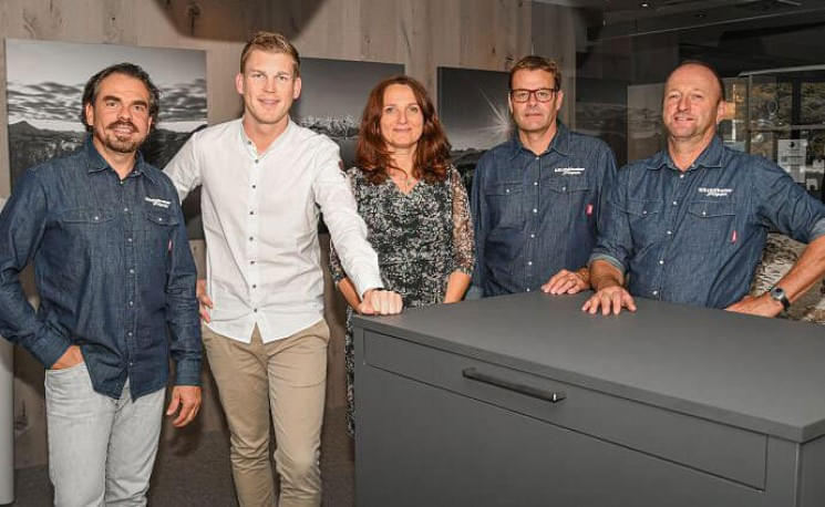 Fünf Personen, drei Männer in blauen Hemden und zwei in gemusterten Hemden, stehen lächelnd um einen Tisch herum in einem modernen Raum.