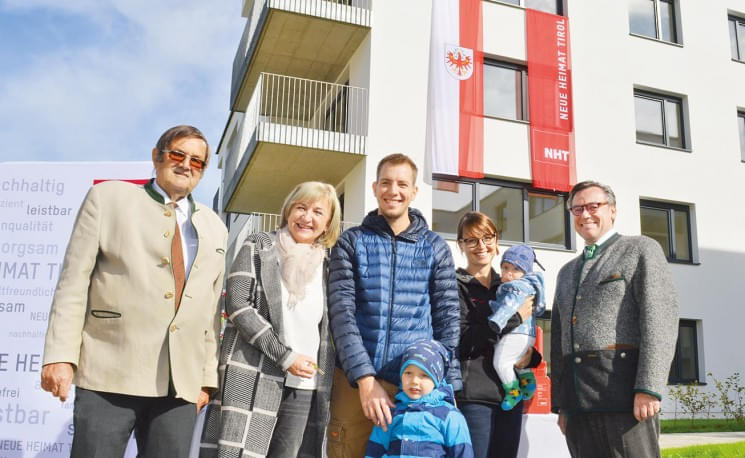 Eine Gruppe von Menschen steht lächelnd vor einem modernen Wohngebäude mit Bannern. Es ist ein sonniger Tag, und die Stimmung scheint festlich zu sein.