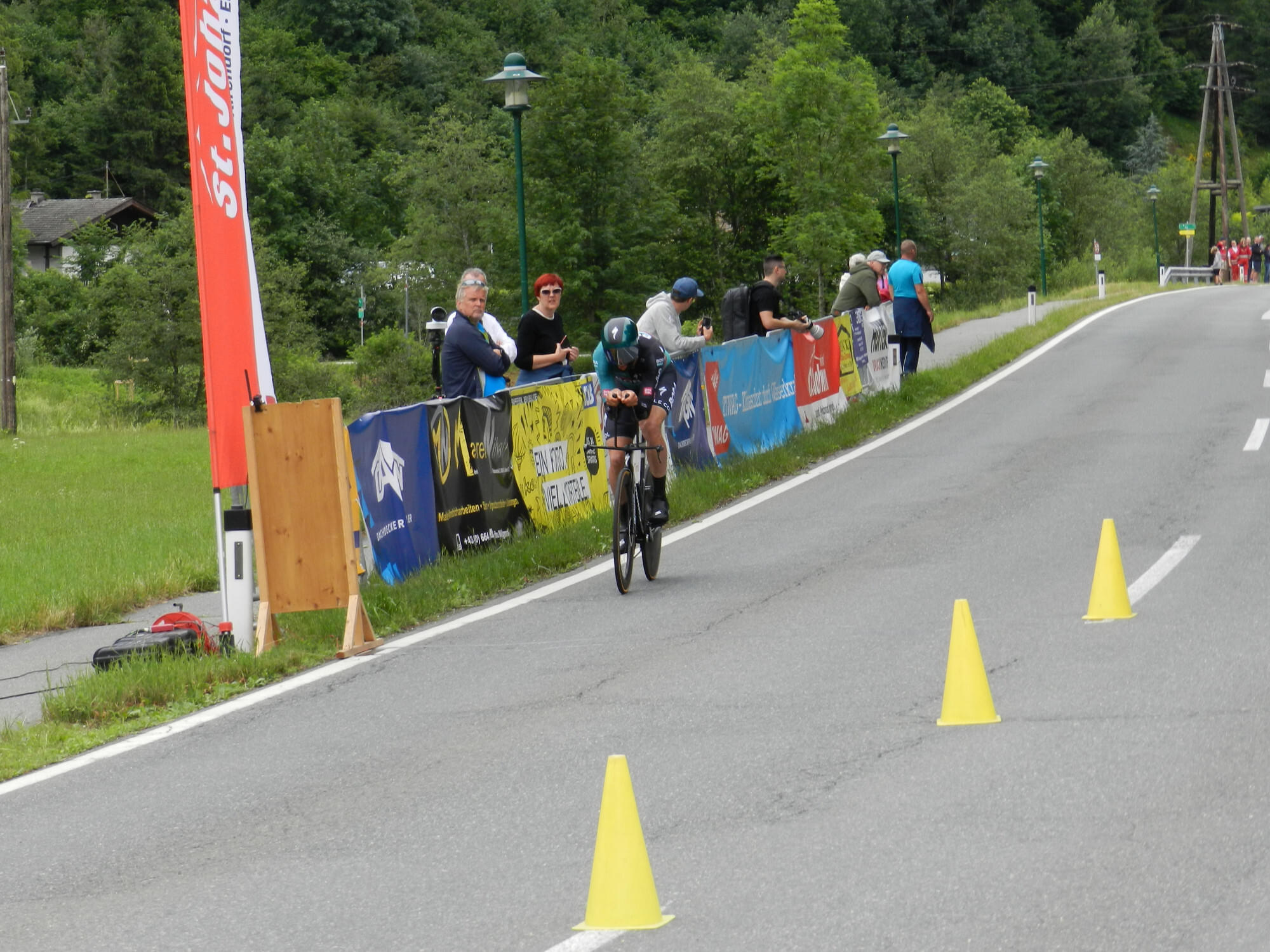 Ein Radfahrer fährt auf einer Landstraße mit gelben Kegeln. Zuschauer stehen am Straßenrand neben bunten Bannern und grüner Landschaft.