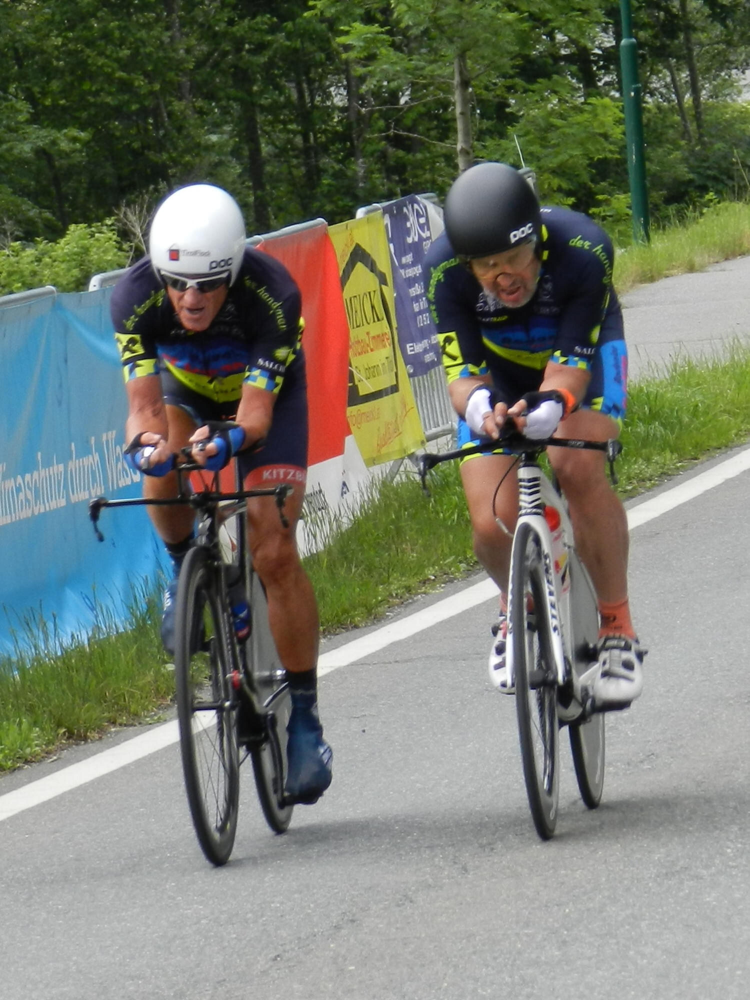 Zwei Radrennfahrer in farbigen Trikots fahren mit hoher Geschwindigkeit eine Landstraße entlang. Beide sind in angespannter Rennhaltung vertieft.