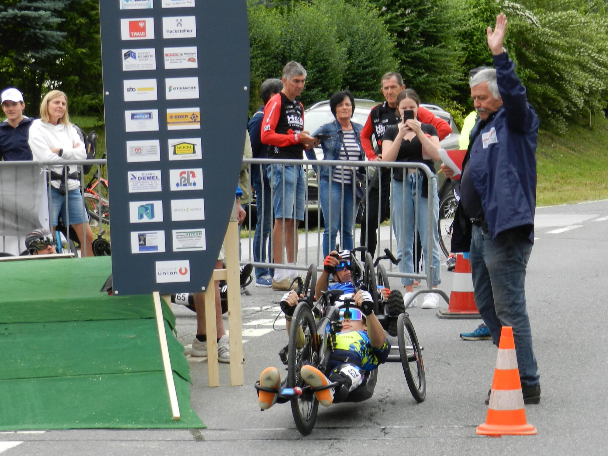 Ein Handbiker überquert die Ziellinie bei einem Rennen, während ein Schiedsrichter mit der Hand winkt. Zuschauer stehen hinter einer Absperrung.