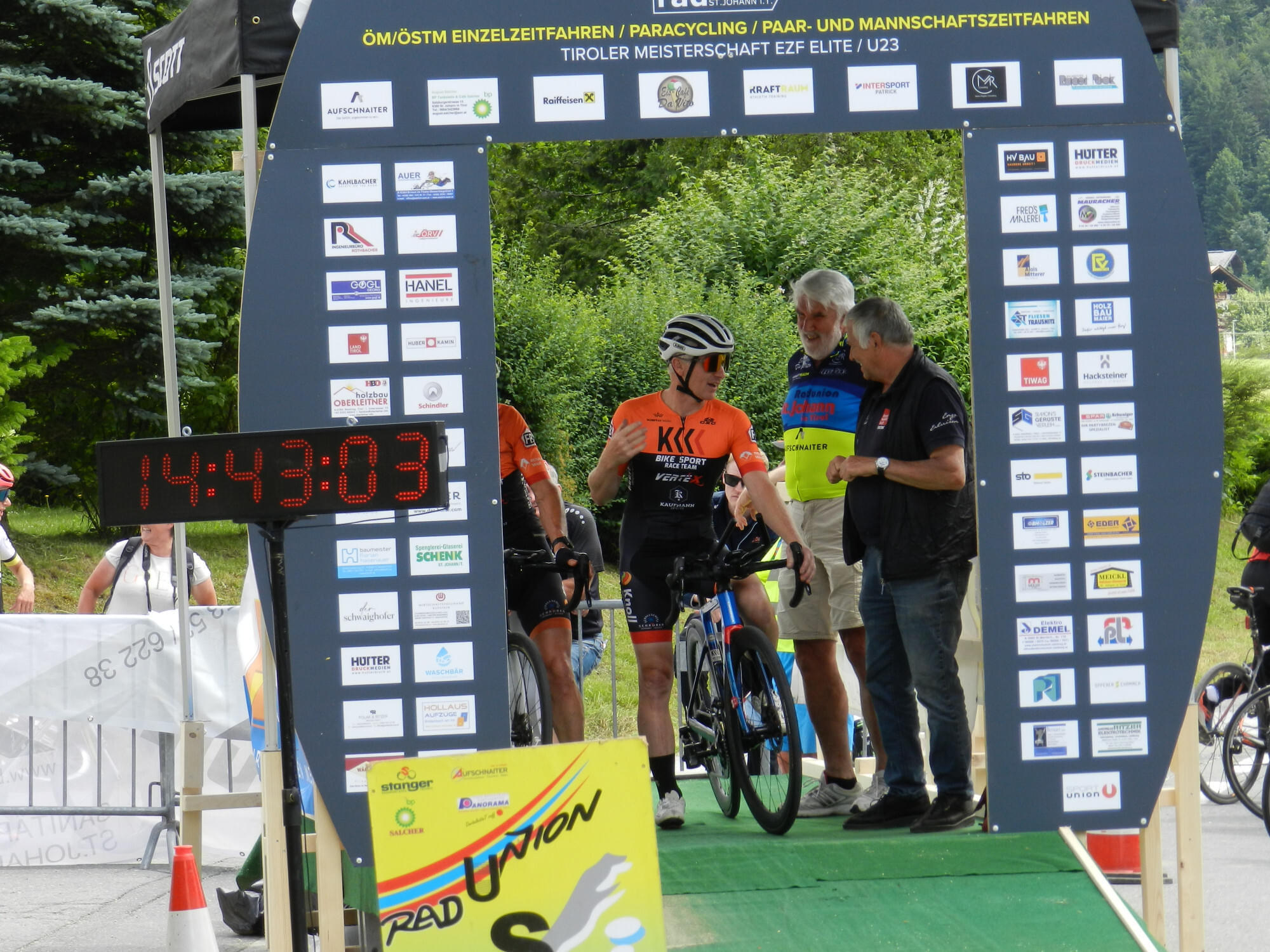 Ein Radfahrer in orangefarbenem Trikot erreicht das Ziel eines Radrennens, umgeben von Helfern und einem großen Timer mit der Zeit 14:49:03.
