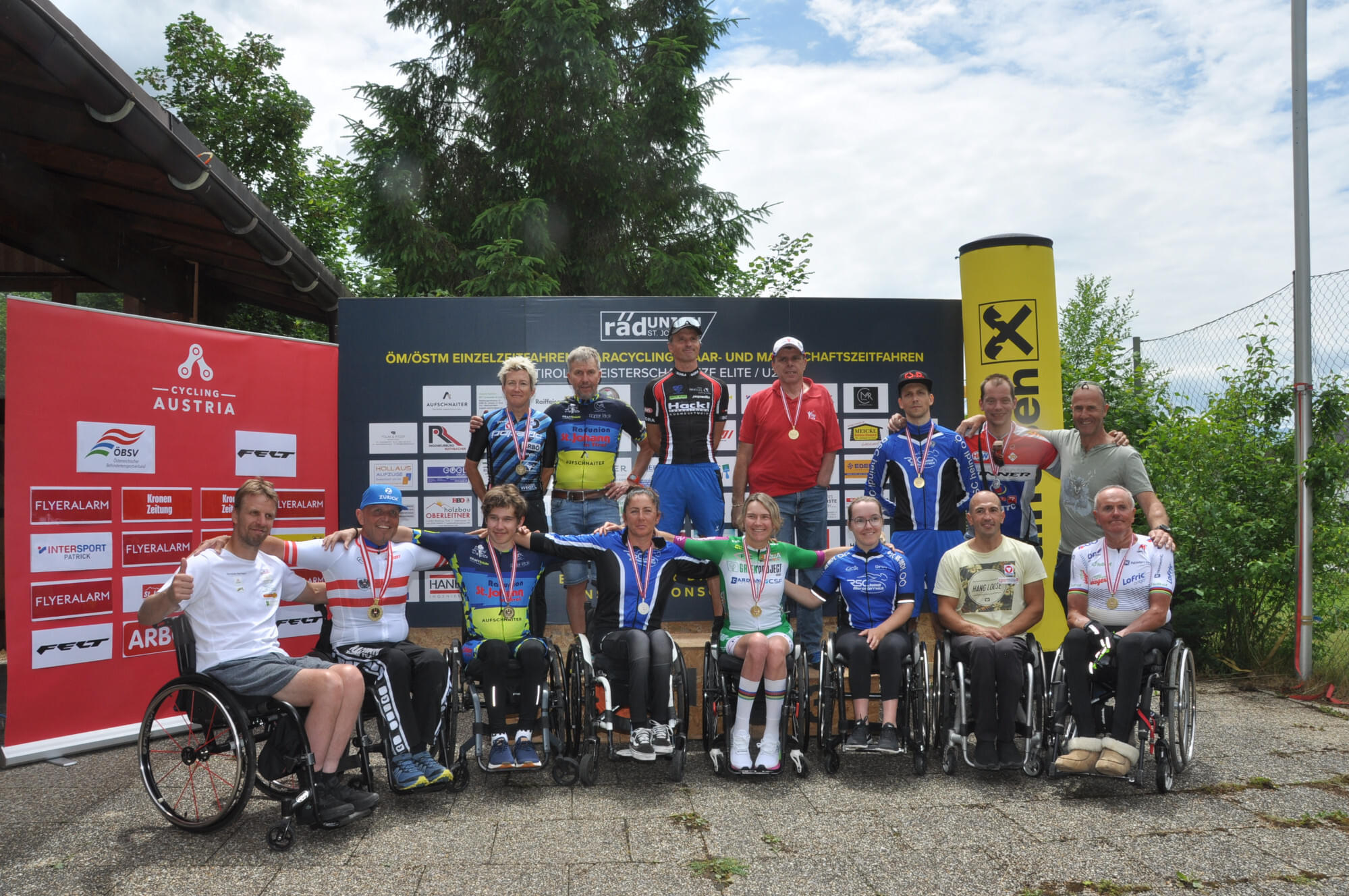 Eine Gruppe von Sportlern in Rollstühlen posiert mit Medaillen vor einem Banner unter freiem Himmel. Sie tragen Sportkleidung, einige haben Trikots an.
