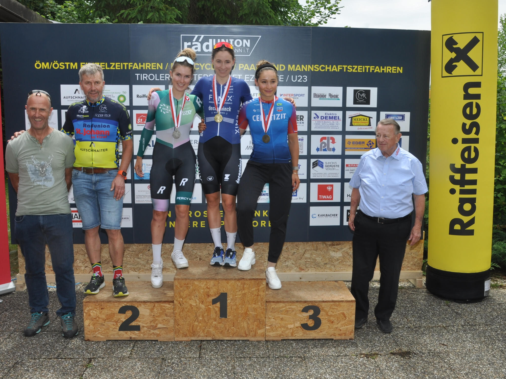 Drei Radrennfahrerinnen stehen mit Medaillen auf einem Podium, flankiert von zwei Offiziellen. Im Hintergrund ist eine Sponsorenwand zu sehen.