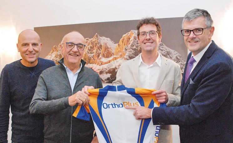 Vier Männer halten gemeinsam ein OrthoPlus-Radtrikot vor einem Bild mit Bergen in einem Raum.