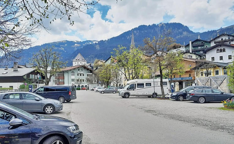 Ein malerischer Dorfplatz mit parkenden Autos, umgeben von traditionellen Gebäuden und Bergen im Hintergrund unter einem bewölkten Himmel.