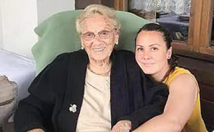 Eine ältere Frau sitzt lächelnd in einem Sessel, während eine jüngere Frau sie liebevoll umarmt. Beide wirken glücklich in einer gemütlichen Umgebung.