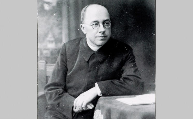 Schwarz-weiß-Foto eines Mannes mit Brille und Anzug, der an einem Tisch sitzt. Er hat kurzes Haar und hält die Hände gefaltet auf dem Tisch.