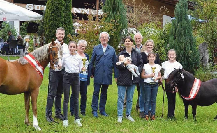 Gruppenfoto im Freien mit acht Personen und zwei Pferden, die Preisschleifen tragen. Ein Baby wird von einer Frau in der Mitte gehalten.