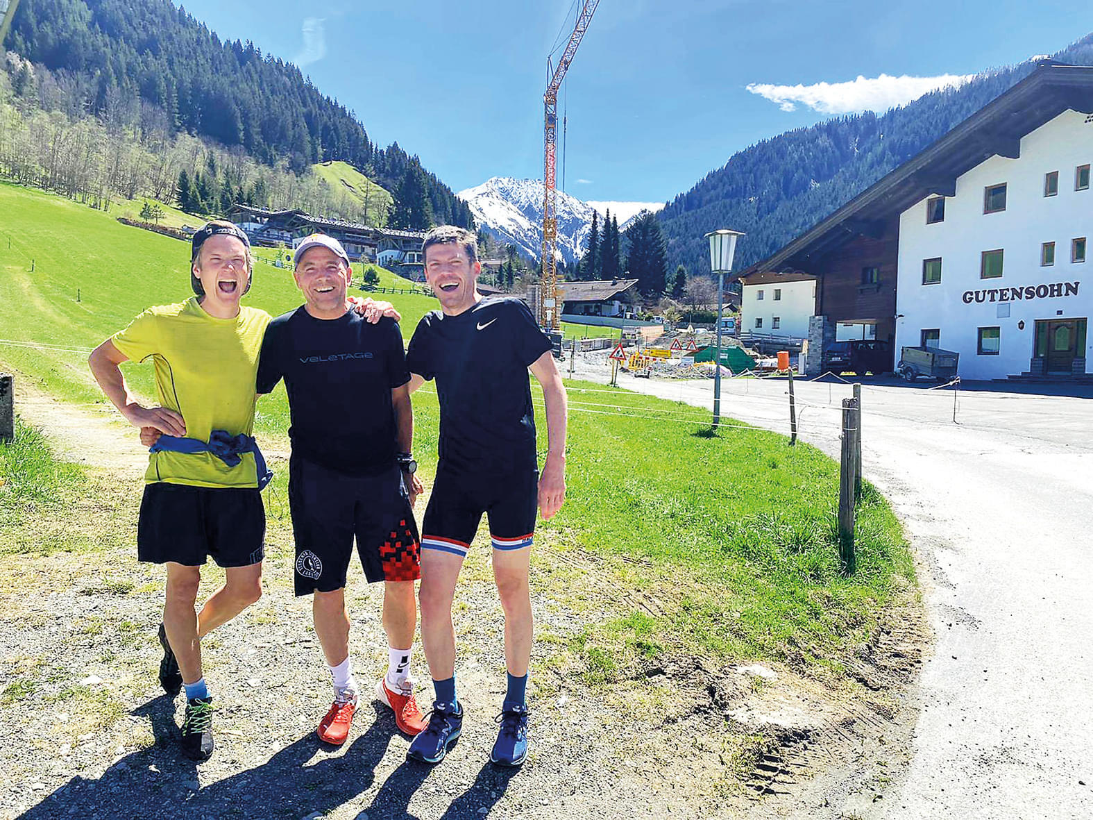 Drei Männer in Sportkleidung stehen lachend vor einer malerischen Bergkulisse. Im Hintergrund sind grüne Wiesen, ein Kran und schneebedeckte Gipfel zu sehen.