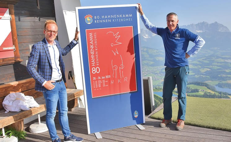 Zwei Männer halten eine Plakattafel mit einem roten und blauen Design, im Hintergrund eine Berglandschaft unter klarem Himmel.