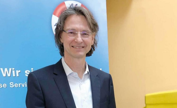 Ein lächelnder Mann mit Brille in einem dunklen Anzug steht vor einem blauen Hintergrund mit weißem Text und einem gelben Element rechts.