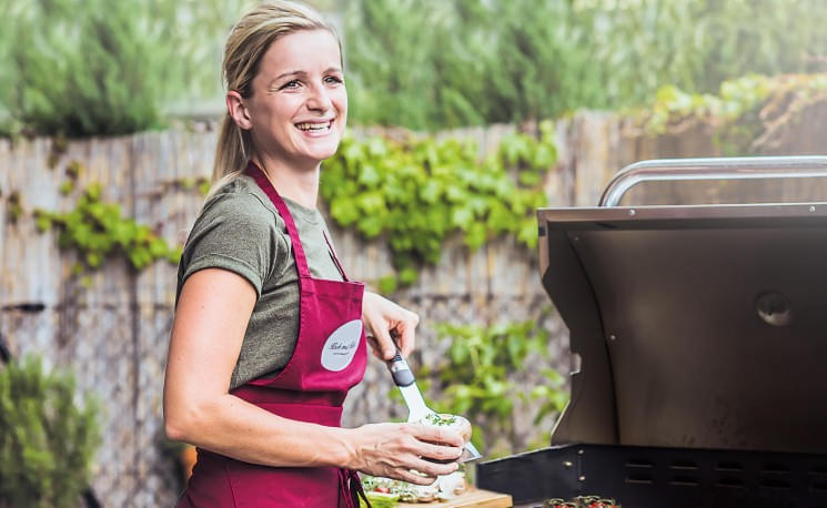 Lächelnde Frau grillt im Garten, trägt rotes Schürzchen, hält Gewürze in der Hand. Im Hintergrund sind Pflanzen und ein Holzzaun zu sehen.