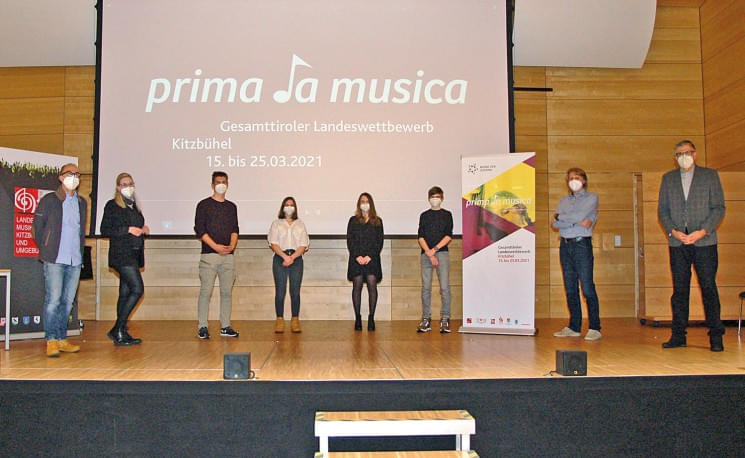 Gruppe von Menschen steht auf einer Bühne vor einem "prima la musica" Banner, alle tragen Masken. Der Wettbewerb fand im März 2021 in Kitzbühel statt.