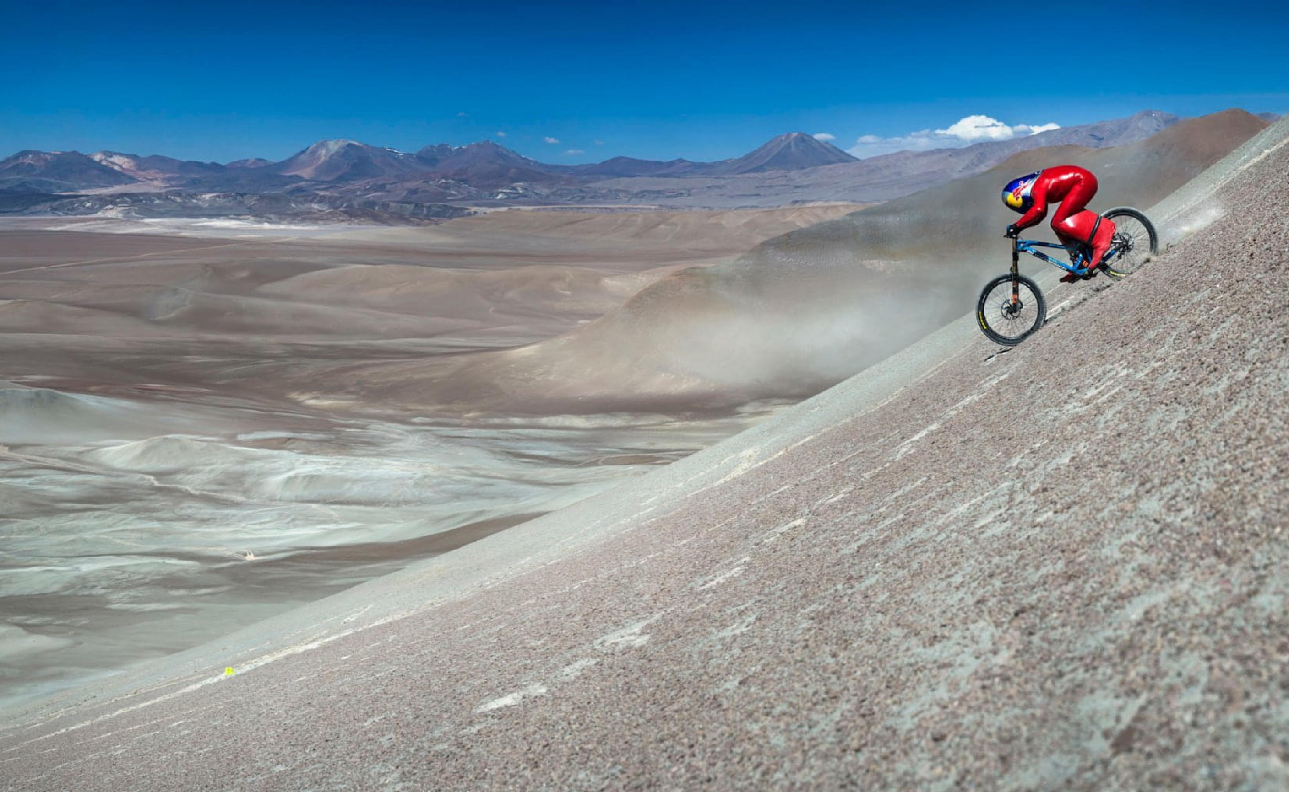 Ein Radfahrer in rotem Anzug fährt mit einem Fahrrad eine steile Sanddüne hinunter, umgeben von einer weitläufigen, kargen Wüstenlandschaft unter klarem blauem Himmel.