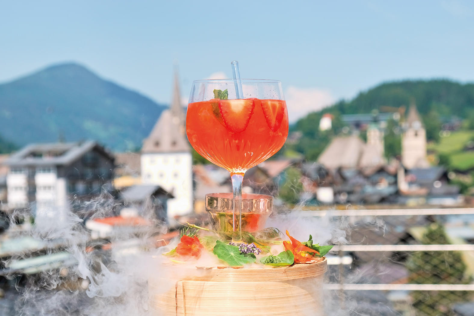 Ein sommerlicher Cocktail auf einem Tisch mit Berglandschaft im Hintergrund. Der Drink ist orange, garniert mit Minze und serviert auf rauchendem Eis.