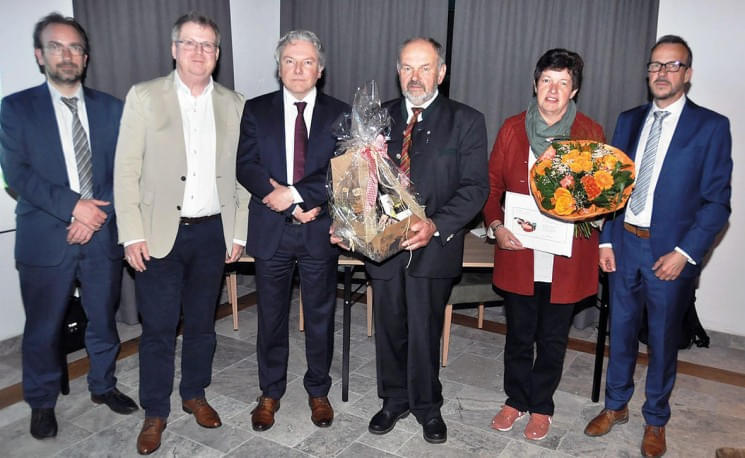 Sechs Personen stehen nebeneinander in formeller Kleidung. Eine Frau hält einen Blumenstrauß, ein Mann ein Geschenkpaket. Hinter ihnen hängen graue Vorhänge.