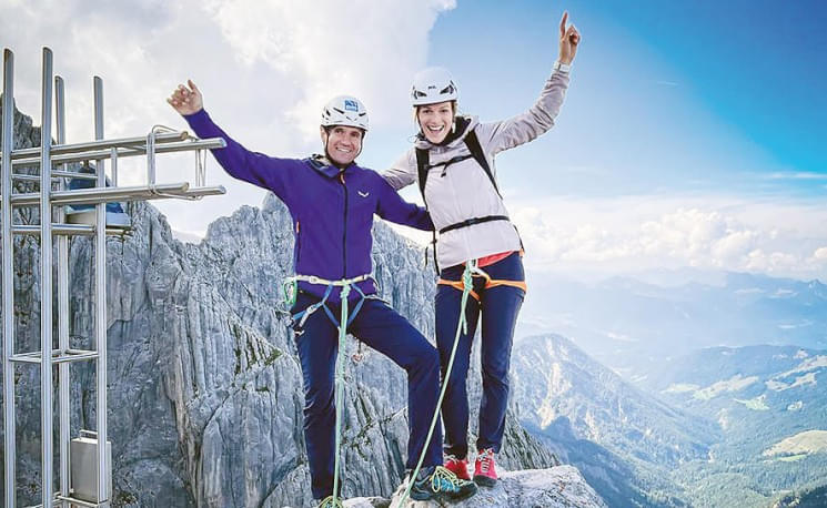 Zwei Personen in Kletterausrüstung stehen auf einem Berggipfel und heben die Arme. Der Himmel ist blau mit ein paar Wolken, umgeben von einem beeindruckenden Bergpanorama.