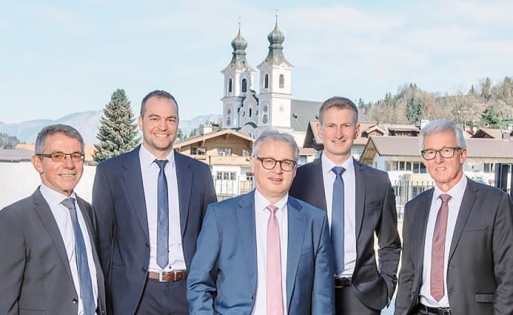 Fünf Männer in Anzügen stehen vor einer idyllischen Kulisse mit Kirchengebäuden und Bergen im Hintergrund, bei klarem, sonnigem Wetter.