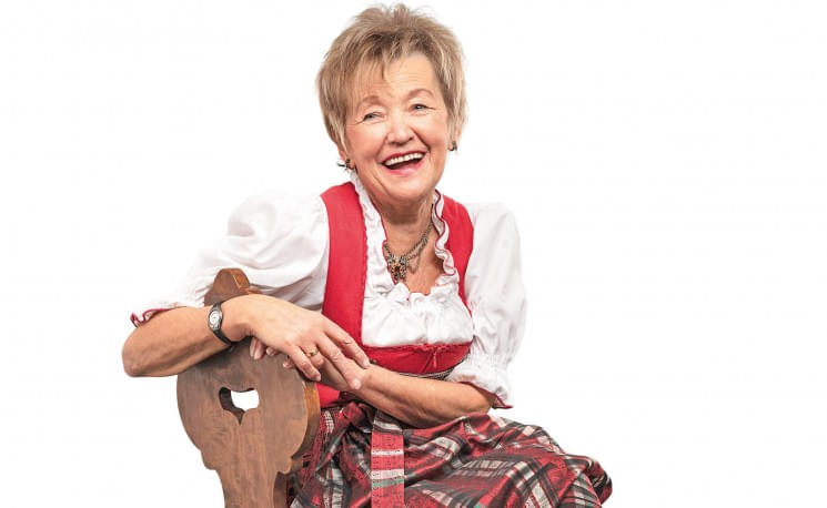 Ältere Frau in traditioneller bayerischer Tracht, lächelnd auf einem Holzstuhl sitzend vor weißem Hintergrund.