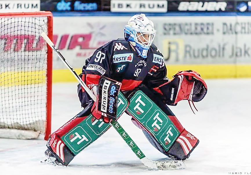 Ein Eishockey-Torwart in voller Ausrüstung steht konzentriert vor dem Tor. Sein Trikot ist dunkel mit Werbeaufdrucken, und er trägt auffällige Schoner.