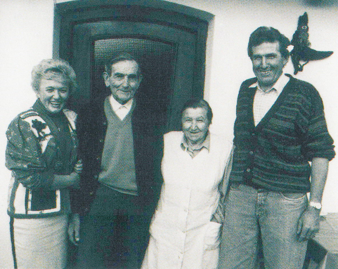 Eine schwarz-weiße Fotografie zeigt vier lächelnde Menschen, zwei Frauen und zwei Männer, die eng beieinander stehen. Im Hintergrund ist eine Tür zu sehen.