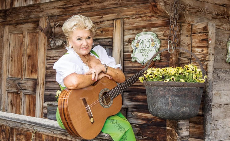 Ältere Frau in traditioneller Tracht lehnt an Holzwand, hält Gitarre. Neben ihr ein Topf mit gelben Blumen und ein Schild mit der Zahl 405.