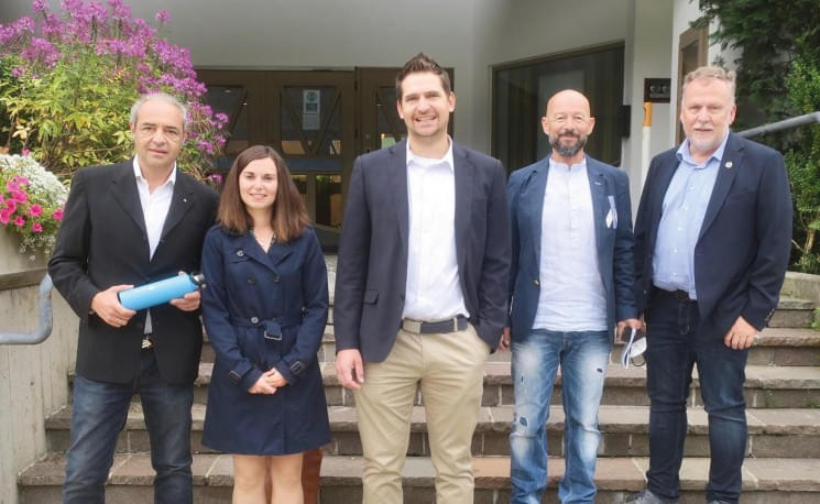 Fünf Personen in formeller Kleidung stehen lächelnd auf einer Treppe vor einem Gebäude. Sie scheinen sich in einer Gruppenbesprechung zu befinden.