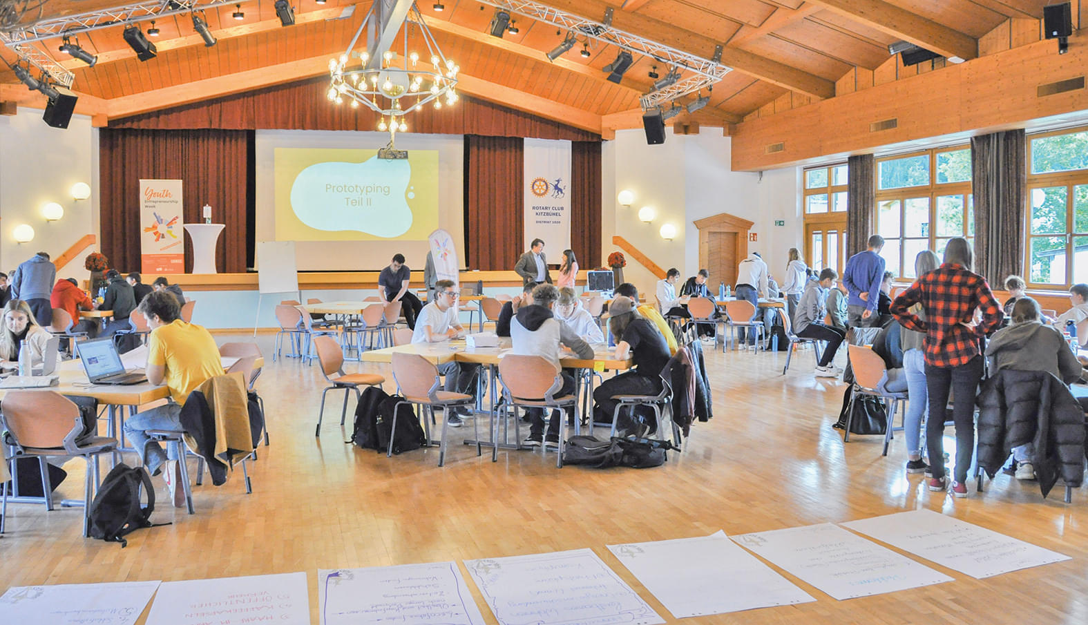 Menschen arbeiten in einem großen, hellen Saal an Tischen mit Laptops. Eine Leinwand zeigt eine Präsentation, und Banner stehen an den Seiten.