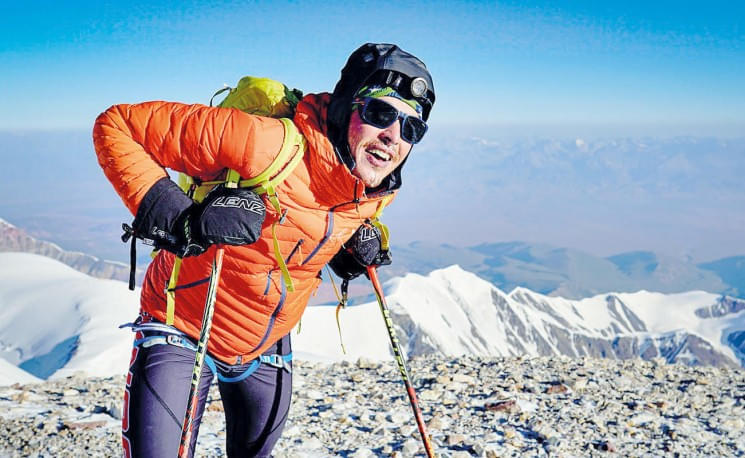 Bergsteiger in heller Kleidung und Helm, mit Skistöcken auf schneebedecktem Gipfel, strahlend unter blauem Himmel. Blick auf entfernte Bergketten im Hintergrund.