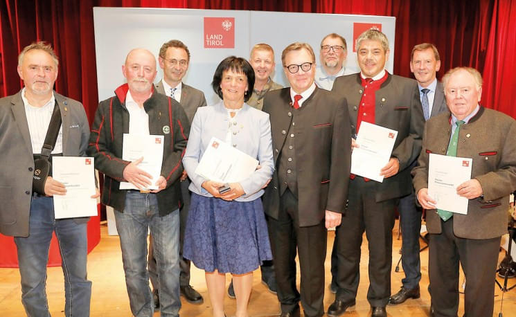 Eine Gruppe von zehn Personen in formeller Kleidung mit Urkunden steht auf einer Bühne vor einem Banner, das "Land Tirol" zeigt.