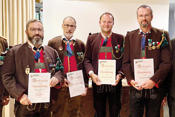 Vier Männer in traditioneller Tiroler Tracht posieren mit Urkunden vor einer weißen Wand, alle lächeln und tragen dekorative Abzeichen.