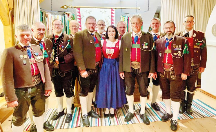 Gruppe von Männern in traditioneller bayerischer Tracht und eine Frau in einem Dirndl lächeln in einem Raum mit Teppichen und Vorhängen.