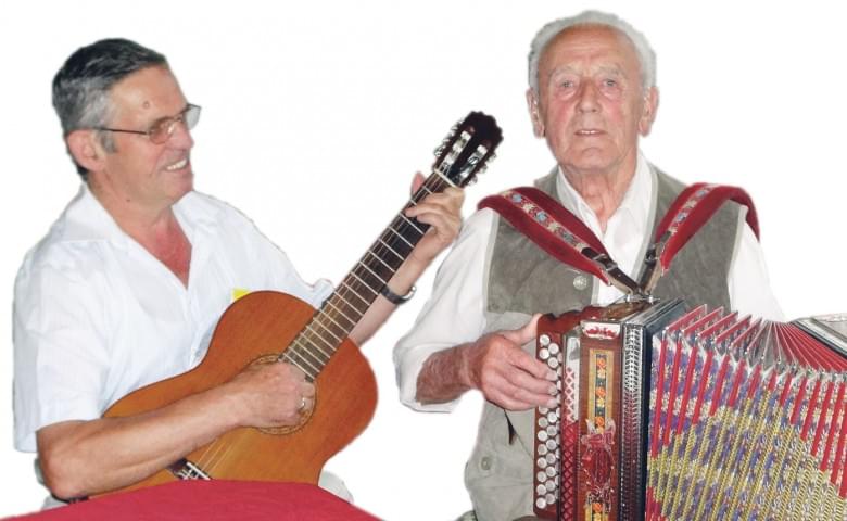 Zwei ältere Männer musizieren gemeinsam. Einer spielt Gitarre, der andere Akkordeon. Beide scheinen konzentriert und erfreuen sich an der Musik.