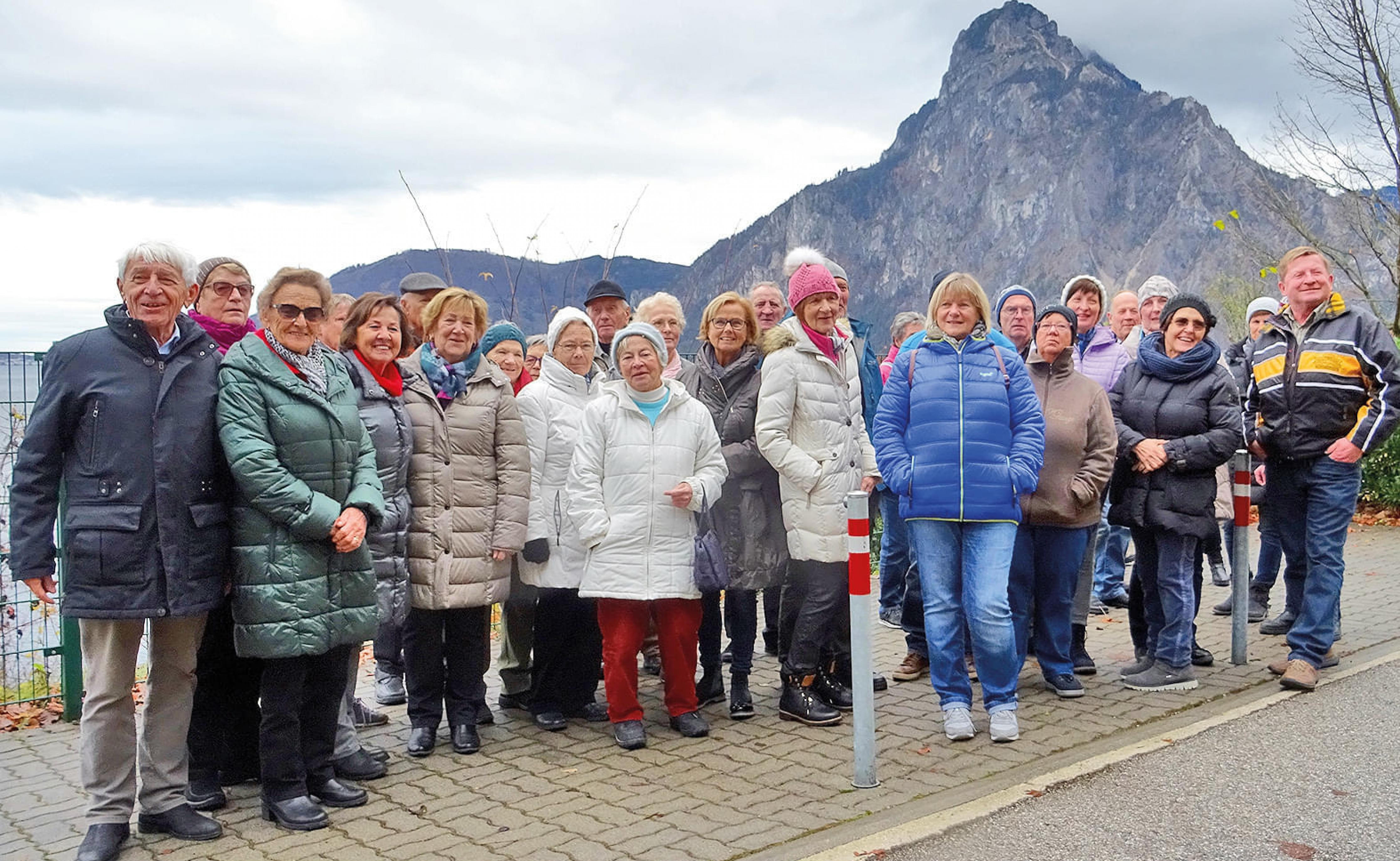 Eine Gruppe von Menschen in Winterkleidung steht vor einer Bergkulisse, lächelt und posiert für ein Foto. Der Himmel ist bewölkt, es scheint kühl zu sein.