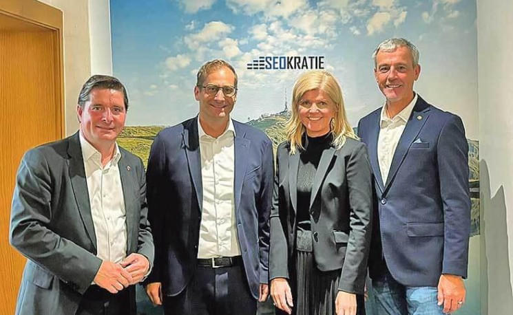Vier Personen in Business-Kleidung stehen vor einem Wandbild mit dem Logo "SEOKRATIE". Alle lächeln in die Kamera, im Hintergrund ist eine grüne Landschaft.