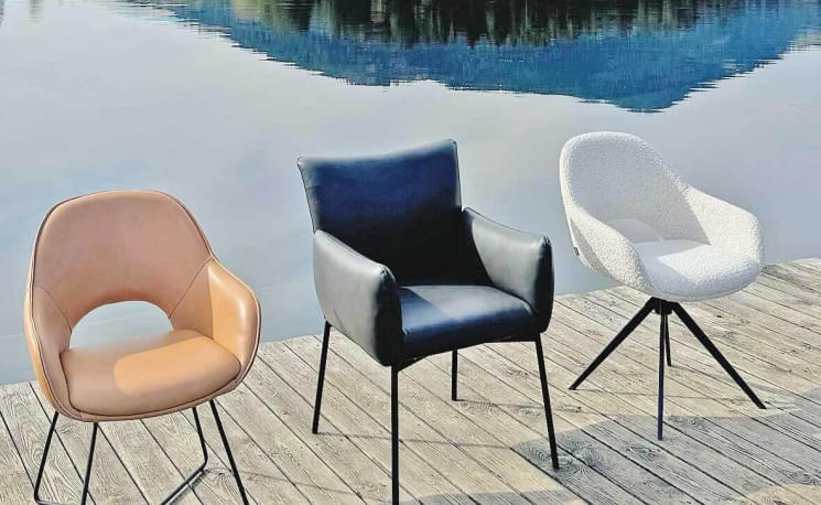 Drei moderne Stühle, in Braun, Schwarz und Weiß, stehen auf einem Holzsteg vor einem ruhigen See mit spiegelndem Wasser und bewaldeten Bergen im Hintergrund.